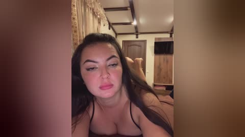 Video of a young Asian woman with long black hair, wearing a black bra, lying on a bed in a dimly lit room, making a pouty face.