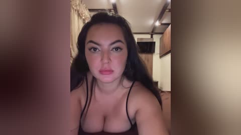 Video of a young, light-skinned woman with long, dark hair, wearing a black spaghetti-strap top, looking directly at the camera in a dimly-lit, ornate room with chandeliers.