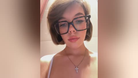Video of a young woman with short, brown hair, wearing large, black-framed glasses and a white spaghetti strap top, against a blurred, peach-colored background. She has fair skin and a small cross necklace.