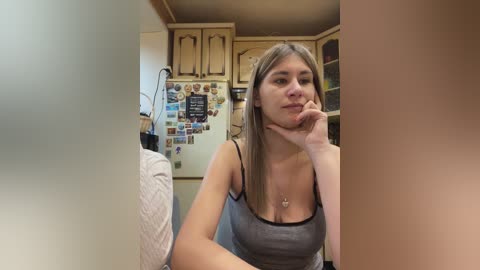 A video of a young woman with light skin, brown hair, and a slender build, wearing a gray tank top, sitting pensively in a cluttered, beige kitchen adorned with magnets and family photos.