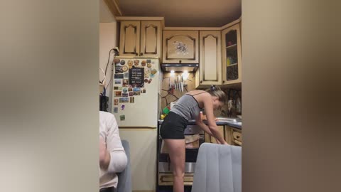 Video of a cozy kitchen with light wooden cabinets, a woman in a gray tank top and black shorts leaning over a sink, and a beige sofa in the background.