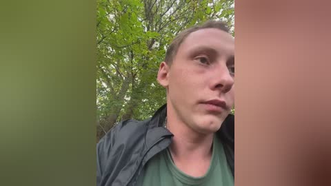A video of a young man with short brown hair, fair skin, and green eyes, wearing a green t-shirt and black jacket, standing in a forest with green trees in the background.