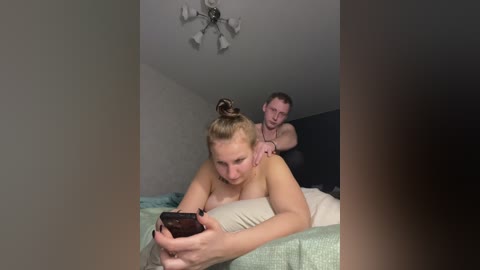 A video of a blonde woman with a bun, topless, lying on a bed, looking at a phone, while a shirtless man massages her back in a dimly lit room.