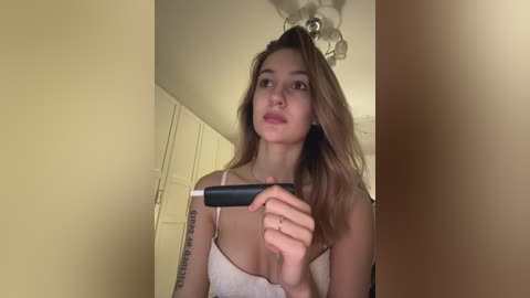 Video of a young woman with long, light brown hair, wearing a white lace camisole, holding a black and silver hair straightener, in a beige-walled room.