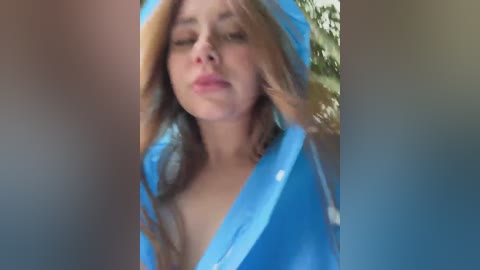 Video of a young woman with long, light brown hair, wearing a blue robe, eyes closed, with a blurred background of greenery.