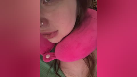 A close-up video of a young woman with fair skin and brown hair, wearing a bright pink fluffy pillow around her neck, partially obscuring her face. The background is blurred and pink-colored.