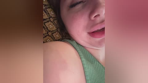 Video of a young woman with light skin and brown hair, partially covered by a green sleeve, lying on a patterned pillow with a yellow and brown design. Her mouth is open, showing teeth and a small amount of drool on her chin.