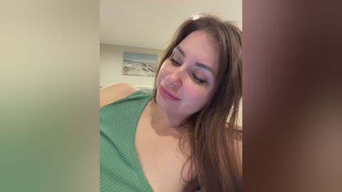 Video of a young woman with light skin and long brown hair, wearing a green ribbed top, smiling gently, standing indoors with blurred background.