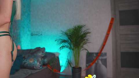 A video showing a woman in green lingerie, standing with her back to the camera, in a dimly lit room with a green-tinted wall, a potted plant, and a patterned pillow on a bed.