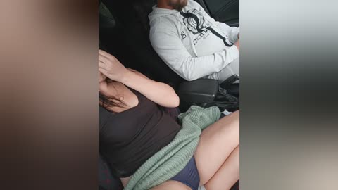Video of a woman with long dark hair, wearing a black top and blue underwear, asleep in a car, with a man in a grey hoodie and black t-shirt sitting next to her.