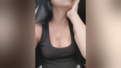 A close-up video of a fair-skinned woman with long, black hair, wearing a sheer black top that reveals her ample cleavage and a delicate gold necklace. Her face is partially obscured by out-of-focus hands.