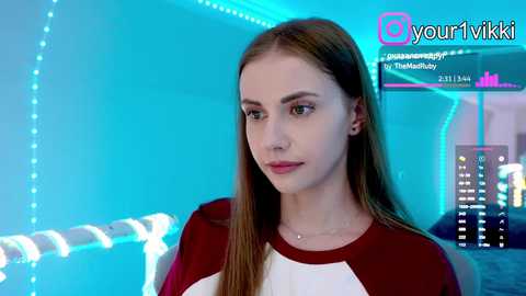 Video of a young woman with long brown hair, wearing a red and white sports top, standing against a blue-lit wall with a digital screen displaying streaming statistics.