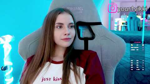 Video of a young Caucasian woman with long brown hair, wearing a red and white sweatshirt, seated in a gray gaming chair. Background features a brightly lit gaming setup with colorful lights and a monitor displaying a chat window.