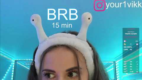 Video of a young woman with fair skin, brown hair, and light makeup, wearing a white headband with two small, white, rabbit-like ears. Background includes a blue and green neon-lit room with a computer screen displaying Twitch chat.