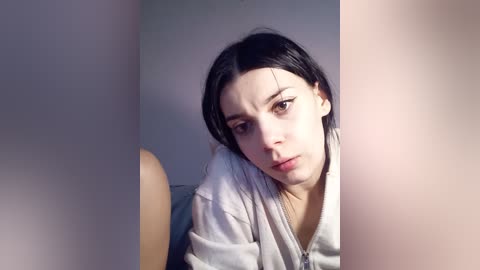 A video of a young woman with pale skin, dark hair, and brown eyes, wearing a gray hoodie, looking directly at the camera with a neutral expression.