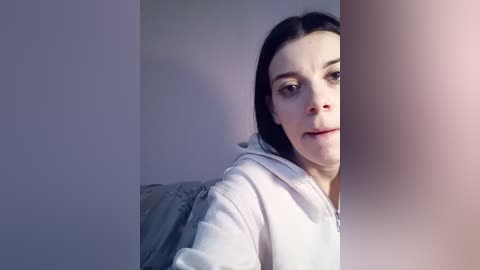 A video of a young woman with light skin, dark hair, and a white hoodie, peeking from behind a large, blurry white pillar, with soft lighting and a minimalist background.