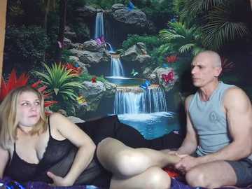 Video of a young blonde woman in a black lace dress and a bald, muscular man in a grey tank top, seated on a bed with a colorful, tropical backdrop featuring a waterfall and jungle.