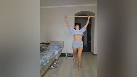 Video of a slender woman in light blue athletic wear, performing a high plank in a minimalist bedroom with light wooden floors, white walls, and an unmade bed.