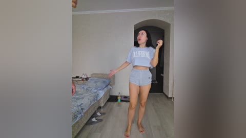 Video of a young woman in a modest, casual outfit, standing in a sparsely furnished bedroom with light wooden floors and white walls. She has long black hair and is looking down, holding a smartphone in her right hand.