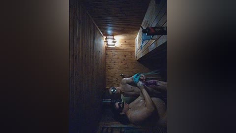 Video of a shirtless, muscular man with wet hair and a blue towel, standing in a dimly lit, narrow wooden shower stall with a light fixture.