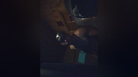 A dimly lit video of a shirtless, muscular man with a helmet, standing in a wooden-paneled room with a fan and a small table.