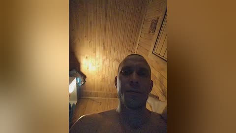 A video of a bald, muscular, shirtless man with a stern expression in a dimly lit, wooden-paneled room. A small lamp illuminates the background, casting a warm glow.