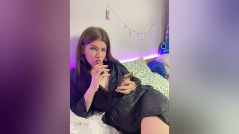 Video of a young woman with long brown hair, wearing a black robe, lying on a bed with floral sheets, holding a phone, looking sultry.