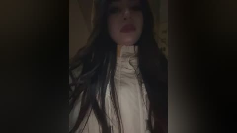 A dimly lit video of a young woman with long black hair, pale skin, and red lips, wearing a white blouse, standing in a dimly lit room with dark walls.