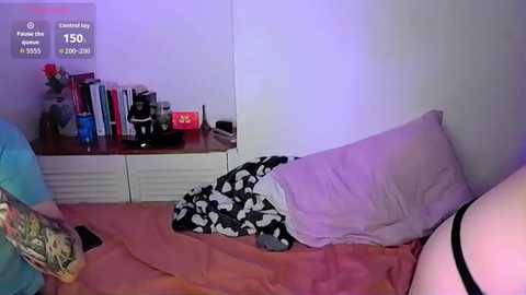 Video of a dimly lit bedroom showing a person in black and white patterned pajamas lying on a pink bedspread. Background includes a wooden nightstand with a clock reading \"15:05,\" a vase of flowers, books, and a black coffee maker.