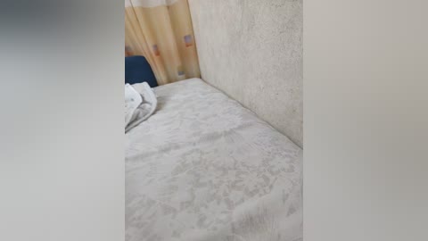 A video of a small, dimly lit room with a white, patterned bedspread, a white pillow, and a mustard-colored curtain partially drawn. The walls are textured and beige, creating a muted, uninviting atmosphere.