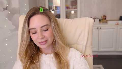 Video of a blonde woman with light skin and long hair, wearing a white sweater, sitting in a beige chair, with a green \"MUA\" watermark on her forehead. Background shows a white cabinet and soft lighting.