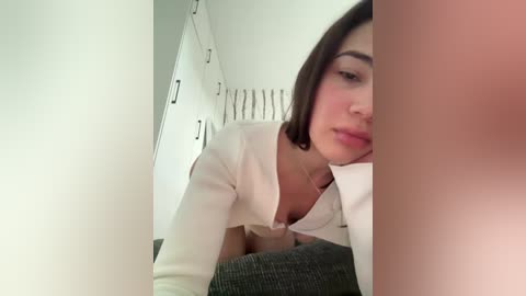 Video of a young woman with light skin and straight dark hair, wearing a white blouse, leaning on a gray couch, in a minimalistic, white-walled room.