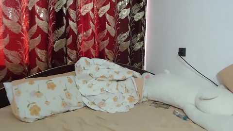 Video of a beige cat sleeping on a bed with white and floral-patterned sheets, next to a dark wooden headboard. Red curtains with gold leaf patterns hang behind the bed, and a white wall with a black electrical outlet is visible.