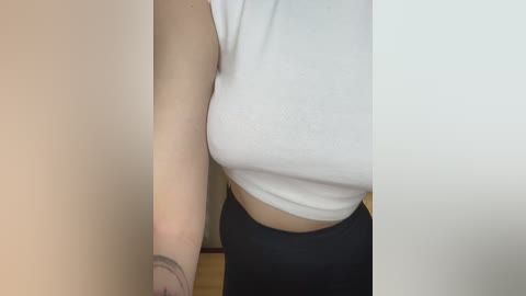 Video of a woman with light skin wearing a tight white crop top that accentuates her large breasts and high-waisted black leggings. She has a tattoo on her forearm.
