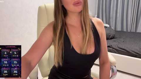 Video of a fair-skinned woman with long blonde hair, wearing a black tank top and tight black pants, sitting in a beige office chair in a modern bedroom with a gray bed and curtains.