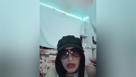 Video of a woman with pale skin, black bob haircut, black sunglasses, and a black beret, wearing red lipstick, standing in a dimly lit room with a tiled floor and white walls.