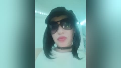 A close-up video of a young woman with pale skin, wearing black sunglasses, a black hat, and a white top. She has a black choker and dark, straight hair. The background is a dimly lit room with soft greenish lighting.