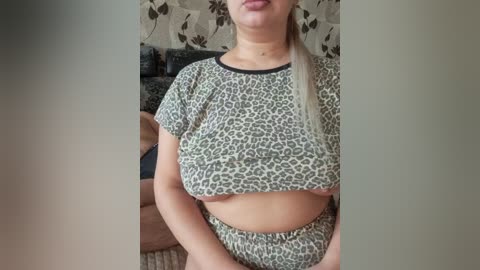 Video of a fair-skinned woman with blonde hair, wearing a leopard-print crop top and matching high-waisted shorts, sitting in a cozy room with floral-patterned wallpaper and a black couch.