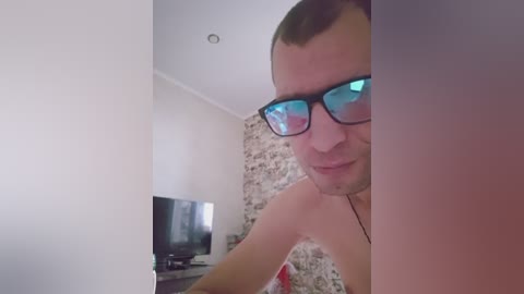 Video of a shirtless man with short brown hair, wearing black rectangular glasses, standing in a modern living room with white walls and a TV on a stand.