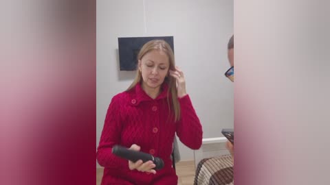 Video of a blonde woman in a red knit coat, adjusting her hair, standing in a minimalist room with a flat-screen TV on the wall.