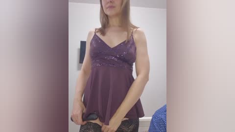 Video of a Caucasian woman with light skin and brown hair, wearing a sheer, purple lace camisole, standing in a minimalist, white-walled room.