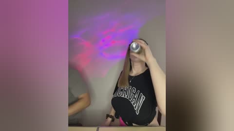 Video of a woman in a black tank top with \"BONJOUR\" text, holding a can, with a purple light beam on a beige wall, blurred background.