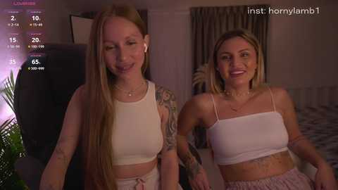 Video of two smiling women with fair skin and blonde hair, wearing crop tops, standing indoors; one with tattoos.