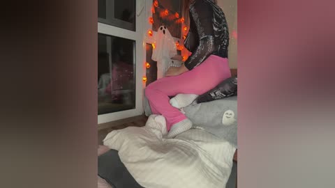 Video of a person with light skin and long hair, wearing a black jacket, pink leggings, and white fuzzy socks, seated on a plush blanket in a cozy, dimly-lit room with orange string lights and a white ghost decoration in the background.