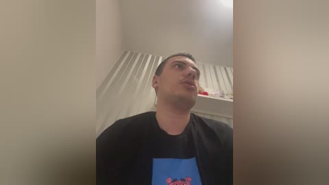 Video of a Caucasian man with short dark hair, wearing a black T-shirt with a blue graphic, standing in a dimly lit room with beige walls and a white radiator.