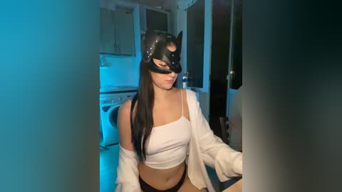 A video of a woman with long black hair, wearing a black cat mask and white crop top, sitting in a dimly lit kitchen with blue lighting.