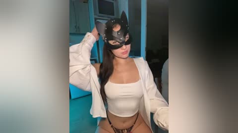 A video of a young woman with light skin and long dark hair, wearing a black cat mask, white cropped top, and black panties, posing seductively indoors.