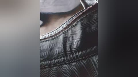 Close-up video of a black, textured, herringbone-patterned leather handbag, focusing on the stitched seams and contrasting metallic zipper. The background is blurred, with soft lighting highlighting the bag's texture and details.