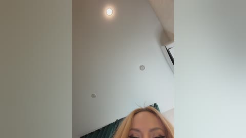 Video of a blonde woman's face, partially obscured by a ceiling light, with a green curtain visible in the background.