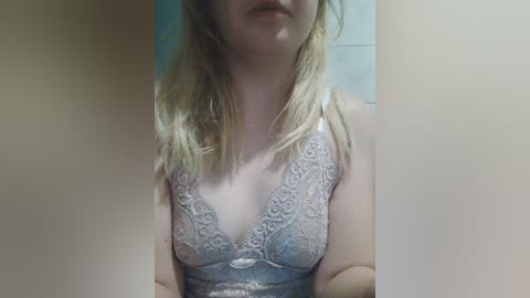 Video of a Caucasian woman with fair skin and shoulder-length blonde hair, wearing a lacy, pale blue bralette. She stands indoors, partially obscured by a beige wall on either side.